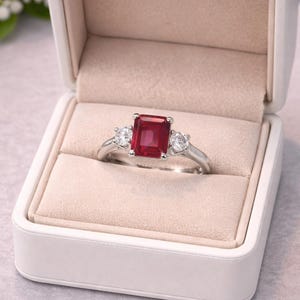 May include: A silver-toned ring featuring a central emerald-cut red gemstone flanked by two smaller round-cut clear stones. The ring is presented inside a white, rectangular, velvet-lined jewelry box with a hinged lid and a soft beige interior.
