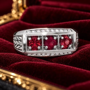 May include: A silver-toned ring featuring three round, deep red gemstones set in a horizontal row. The band is accented with small, clear stones along the top and bottom edges and displays a textured, chevron-like pattern on the side shank.