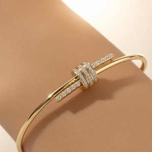 Puede incluir: Una pulsera rígida de oro con un diseño de nudo central adornado con pequeñas piedras claras y brillantes. La banda de metal es lisa y pulida, y el elemento del nudo se extiende en dos barras cortas y rectas incrustadas con piedras.