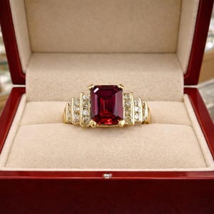 May include: A gold ring featuring a large, rectangular, deep red gemstone. The ring is adorned with smaller, clear gemstones flanking the central stone. It is presented in a red jewellery box with a cream-coloured interior.