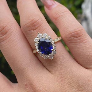 May include: A gold ring featuring a large, oval-cut, deep blue gemstone surrounded by smaller, clear gemstones. The ring's band is also gold. The ring is on a person's finger, with a blurred green background.