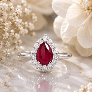 May include: A pear-shaped red gemstone ring set in a polished silver-toned metal band. The central red stone is surrounded by a halo of small, round, clear white stones, creating a classic floral-inspired design with a reflective metallic finish.