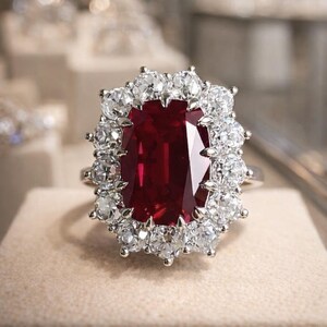 Puede incluir: Un anillo que presenta una gran piedra preciosa rectangular de color rojo intenso en el centro, rodeada por un halo de doce piedras blancas redondas, claras y facetadas engastadas en una banda metálica. El diseño resalta el contraste entre la piedra roja central y las piedras blancas circundantes.