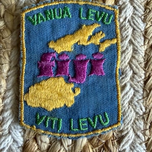 May include: A rectangular embroidered patch with a blue fabric background and a yellow border. It features green text reading VANUA LEVU at the top and VITI LEVU at the bottom. The center displays yellow island shapes and the purple text FIJI.