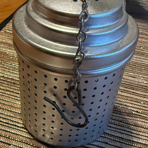 Puede incluir: Un infusor de té cilíndrico de metal plateado con cuerpo perforado y tapa a juego. Una fina cadena de metal está unida a la parte superior de la tapa y termina en un gancho de metal oscuro para asegurar el infusor al borde de un recipiente.