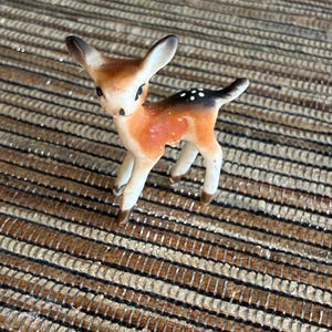 May include: A small ceramic figurine of a fawn with a light brown and white coat, featuring dark brown ears, a dark brown tail, and white spots on its back. The deer stands on four legs with a smooth, glossy finish and painted facial details.