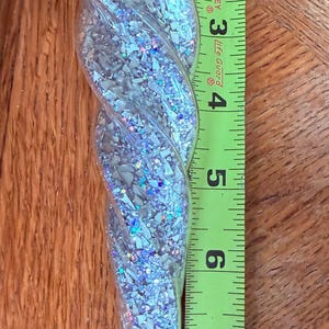 May include: A clear, spiral-shaped glass icicle ornament filled with iridescent silver glitter shards. A yellow Stanley measuring tape placed alongside shows the ornament measures approximately 7.5 inches in total length from top to tip.