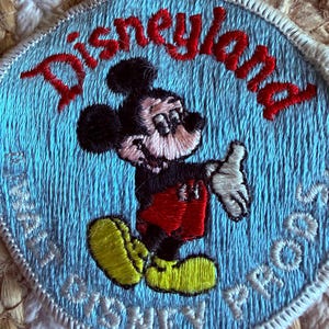 Könnte beinhalten: Ein kreisförmiger gestickter Aufnäher mit hellblauem Hintergrund und dem roten Schriftzug Disneyland in einem Bogen oben. In der Mitte ist Mickey Mouse mit schwarzem Fell, roter Hose und gelben Schuhen abgebildet. Unten steht Disney Props.