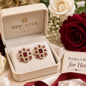 Puede incluir: Un par de pendientes dorados adornados, con piedras centrales rojas y cristales transparentes, presentados en un joyero beige abierto. El conjunto se encuentra frente a rosas rojas y blancas, con una tarjeta que dice "Perfect Gift for Her".