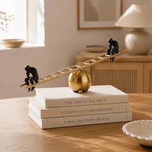 Pode incluir: Uma escultura decorativa com duas figuras humanas pretas sentadas nas extremidades opostas de uma escada dourada equilibrada sobre uma esfera dourada. A escultura repousa sobre uma pilha de três livros intitulados The Art of Balance, Scandinavian Modernism e Form and Proportion.