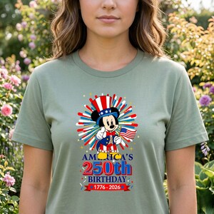 May include: Sage green t-shirt featuring a patriotic Mickey Mouse design. Mickey wears a top hat and holds an American flag, with text that reads "America's 250th Birthday 1776-2026". The design includes red, white, and blue fireworks.