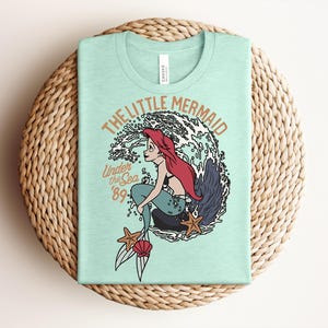 May include: A light mint green t-shirt featuring a graphic of a red-haired mermaid sitting on a rock in front of a large wave. The text reads THE LITTLE MERMAID, Under the Sea, and '89. Two starfish and a pink seashell are at the bottom.