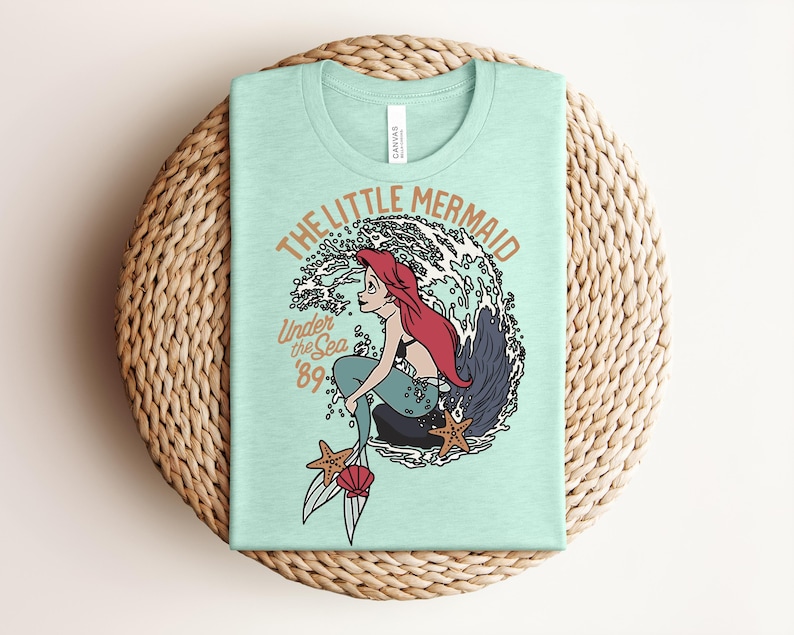 May include: A light mint green t-shirt featuring a graphic of a red-haired mermaid sitting on a rock in front of a large wave. The text reads THE LITTLE MERMAID, Under the Sea, and '89. Two starfish and a pink seashell are at the bottom.