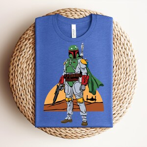 May include: Blue t-shirt featuring a graphic of a character in green and gray armor, holding a weapon, with a green cape. The design includes a desert landscape and a setting sun. The shirt is folded on a woven surface.