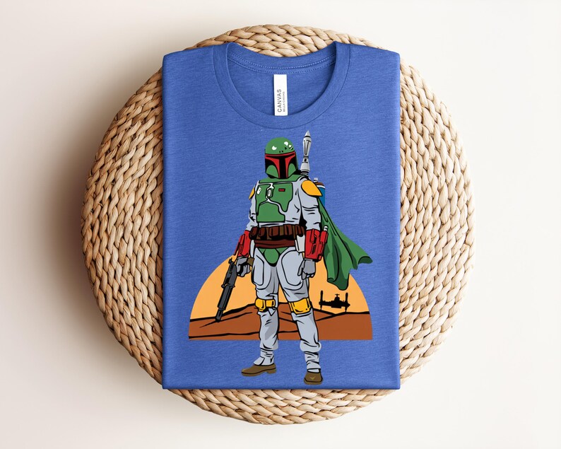 May include: Blue t-shirt featuring a graphic of a character in green and gray armor, holding a weapon, with a green cape. The design includes a desert landscape and a setting sun. The shirt is folded on a woven surface.