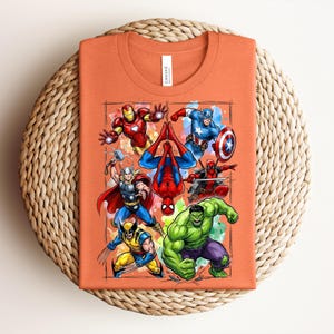 May include: Orange t-shirt with a vibrant graphic of Marvel superheroes such as Iron Man, Captain America, and Spider-Man. The shirt is folded and displayed on a circular woven mat.