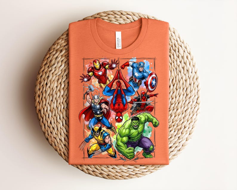 May include: Orange t-shirt with a vibrant graphic of Marvel superheroes such as Iron Man, Captain America, and Spider-Man. The shirt is folded and displayed on a circular woven mat.