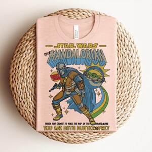 Puede incluir: Una camiseta rosa claro con un gr&aacute;fico de The Mandalorian y Grogu. El texto dice Star Wars The Mandalorian, Bounty Hunter y When You Choose To Walk The Way Of The Mandalore You Are Both Hunter & Prey. El dise&ntilde;o utiliza colores azul, amarillo y verde.