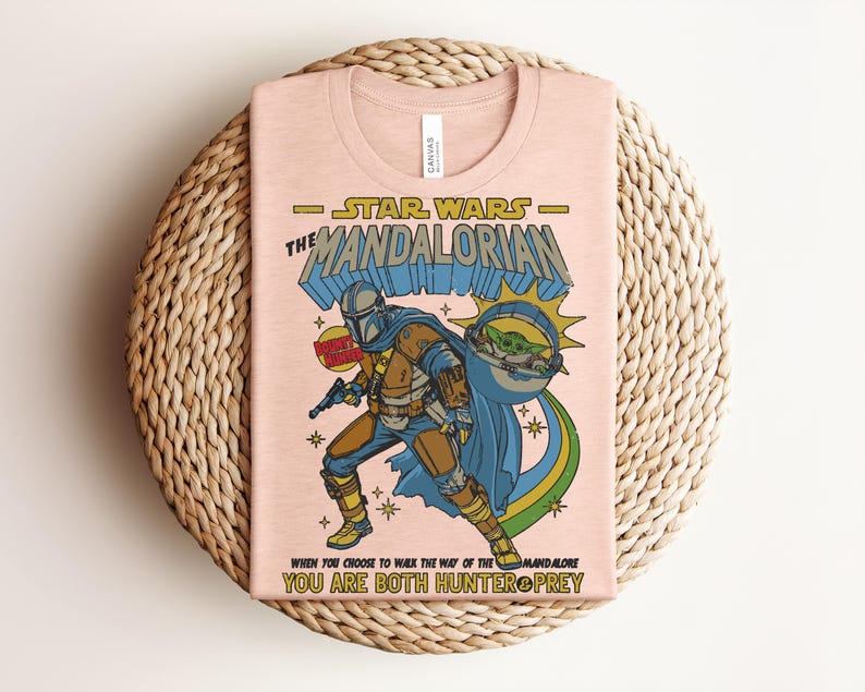 Puede incluir: Una camiseta rosa claro con un gr&aacute;fico de The Mandalorian y Grogu. El texto dice Star Wars The Mandalorian, Bounty Hunter y When You Choose To Walk The Way Of The Mandalore You Are Both Hunter & Prey. El dise&ntilde;o utiliza colores azul, amarillo y verde.