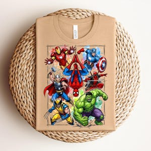 May include: A beige t-shirt with a vibrant illustration of Marvel superheroes, such as Iron Man, Captain America, Spider-Man, Deadpool, Hulk, Thor, and Wolverine. The design is framed with a watercolour-style border.