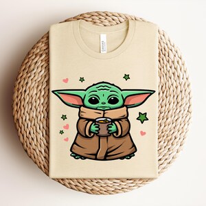 May include: Beige t-shirt featuring a cartoon illustration of a green alien character holding a cup. The character is wearing a brown cloak and has large ears. Pink hearts and green stars surround the character.