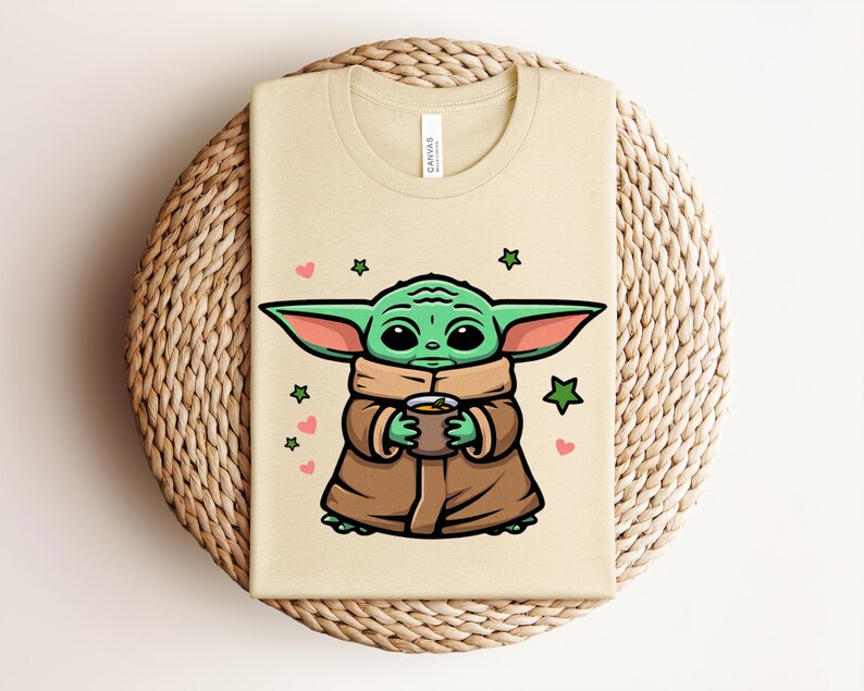 May include: Beige t-shirt featuring a cartoon illustration of a green alien character holding a cup. The character is wearing a brown cloak and has large ears. Pink hearts and green stars surround the character.