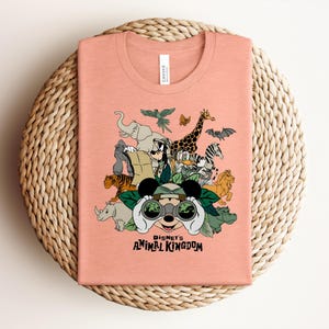 Puede incluir: Una camiseta color melocot&oacute;n con un estampado gr&aacute;fico de personajes de Disney y varios animales, incluyendo un elefante, una jirafa, un tigre y una cebra. El dise&ntilde;o incluye el texto Disney's ANIMAL KINGDOM y muestra a un personaje con binoculares.
