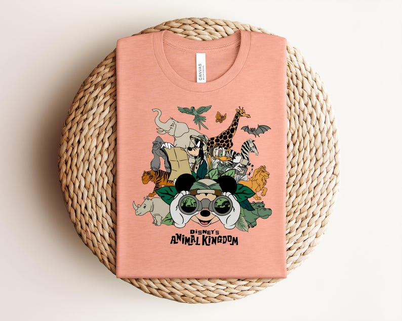 Puede incluir: Una camiseta color melocot&oacute;n con un estampado gr&aacute;fico de personajes de Disney y varios animales, incluyendo un elefante, una jirafa, un tigre y una cebra. El dise&ntilde;o incluye el texto Disney's ANIMAL KINGDOM y muestra a un personaje con binoculares.