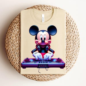 May include: Beige t-shirt featuring a vibrant graphic of Mickey Mouse as a DJ. Mickey is wearing headphones and operating a purple and blue DJ mixer. The shirt is folded on a woven, circular surface.