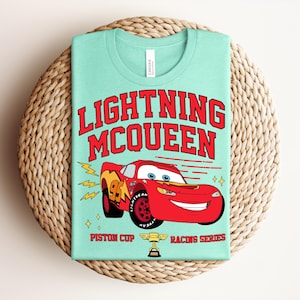 May include: A light green t-shirt featuring Lightning McQueen from the movie Cars. The red graphic includes the text "LIGHTNING McQUEEN" and an image of the race car. Below the car are the words "Piston Cup Racing Series".