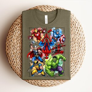 May include: Olive green t-shirt with a vibrant graphic of superheroes such as Iron Man, Captain America, Spider-Man, Thor, Wolverine, and the Hulk. The folded shirt rests on a woven, circular mat.