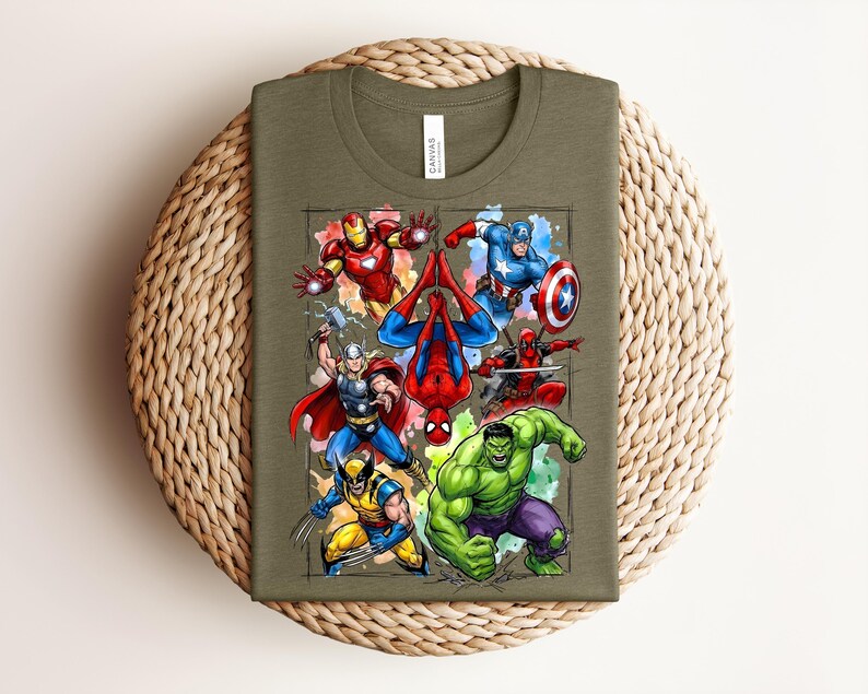 May include: Olive green t-shirt with a vibrant graphic of superheroes such as Iron Man, Captain America, Spider-Man, Thor, Wolverine, and the Hulk. The folded shirt rests on a woven, circular mat.