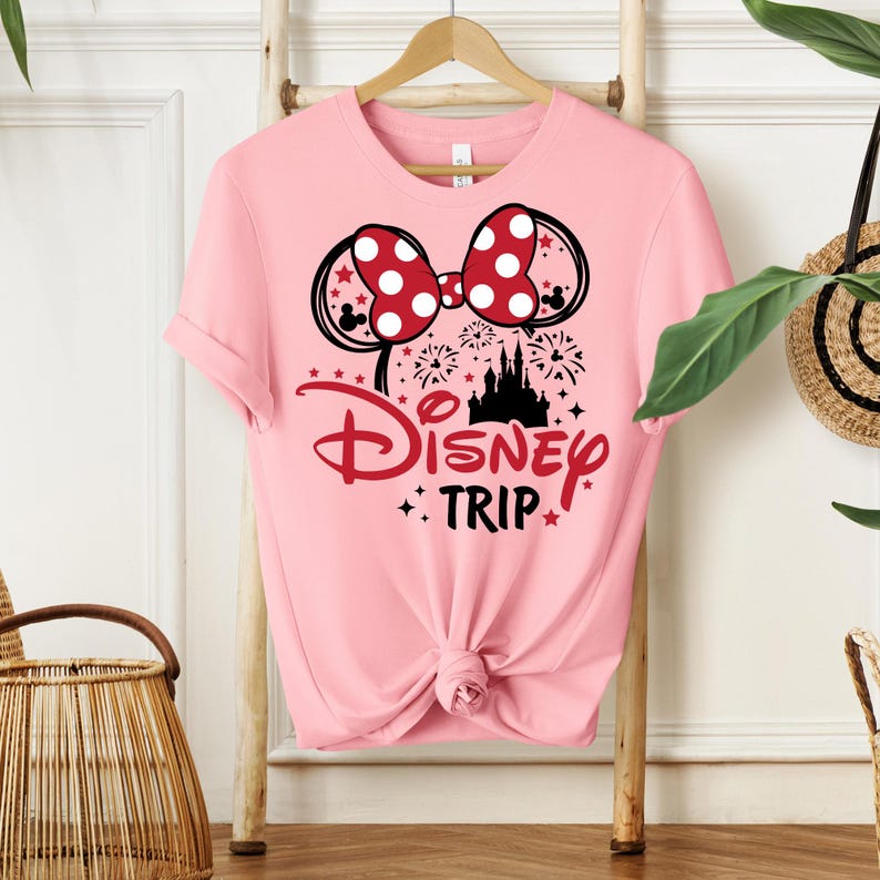 May include: A light pink t-shirt featuring a Minnie Mouse bow with white polka dots and black mouse ears. The shirt has the words "Disney Trip" in red script and a black castle graphic.
