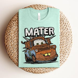 May include: A light mint green crew neck t-shirt featuring a graphic illustration of the character Mater from the movie Cars. The design includes the name MATER in large white letters above the rusty brown tow truck with green eyes and teeth.
