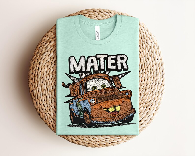 May include: A light mint green crew neck t-shirt featuring a graphic illustration of the character Mater from the movie Cars. The design includes the name MATER in large white letters above the rusty brown tow truck with green eyes and teeth.