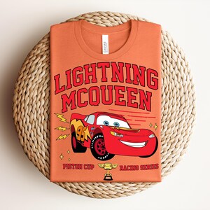 May include: Orange t-shirt featuring Lightning McQueen from the movie Cars. The design includes the text "LIGHTNING MCQUEEN" in red, a red race car, and the words "Piston Cup Racing Series".