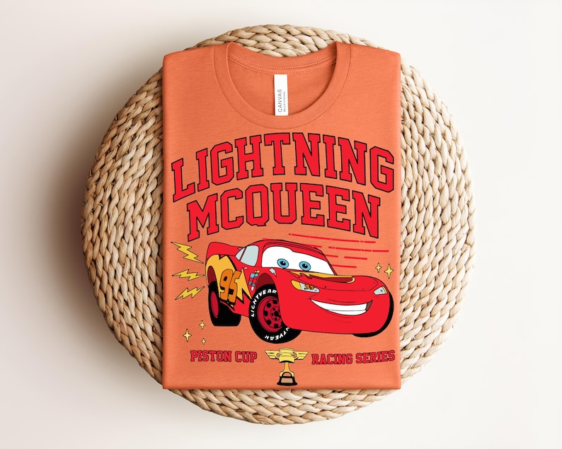 May include: Orange t-shirt featuring Lightning McQueen from the movie Cars. The design includes the text "LIGHTNING MCQUEEN" in red, a red race car, and the words "Piston Cup Racing Series".