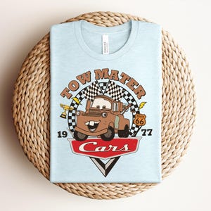 May include: A light blue t-shirt featuring a graphic of the character Tow Mater from the movie Cars. The design includes a checkered flag, a gold trophy, a Route 66 sign, the text Tow Mater, 1977, and the Cars logo in red and white.