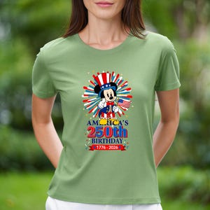 May include: A light green t-shirt featuring a graphic of Mickey Mouse in patriotic attire, holding a small American flag. The shirt celebrates America's 250th birthday, with text reading "AMERICA'S 250th BIRTHDAY 1776-2026."