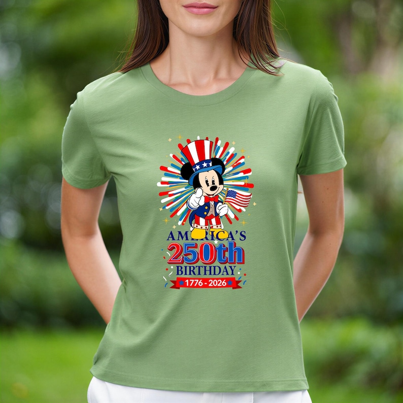 May include: A light green t-shirt featuring a graphic of Mickey Mouse in patriotic attire, holding a small American flag. The shirt celebrates America's 250th birthday, with text reading "AMERICA'S 250th BIRTHDAY 1776-2026."