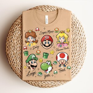 May include: Tan t-shirt featuring colorful illustrations of Super Mario characters, including Mario, Luigi, Princess Peach, Daisy, Yoshi, and Toad. The words "Super Mario" and character names are also printed on the shirt. The shirt is folded on a woven mat.