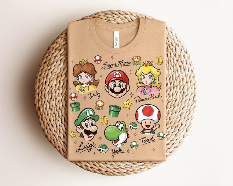 May include: Tan t-shirt featuring colorful illustrations of Super Mario characters, including Mario, Luigi, Princess Peach, Daisy, Yoshi, and Toad. The words "Super Mario" and character names are also printed on the shirt. The shirt is folded on a woven mat.