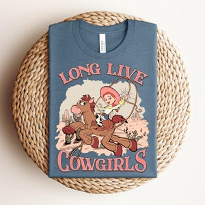 May include: A blue t-shirt featuring a graphic of a cartoon cowgirl riding a brown horse while holding a lasso. The text "LONG LIVE COWGIRLS" is printed in pink above and below the illustration, which includes cacti and a desert landscape.