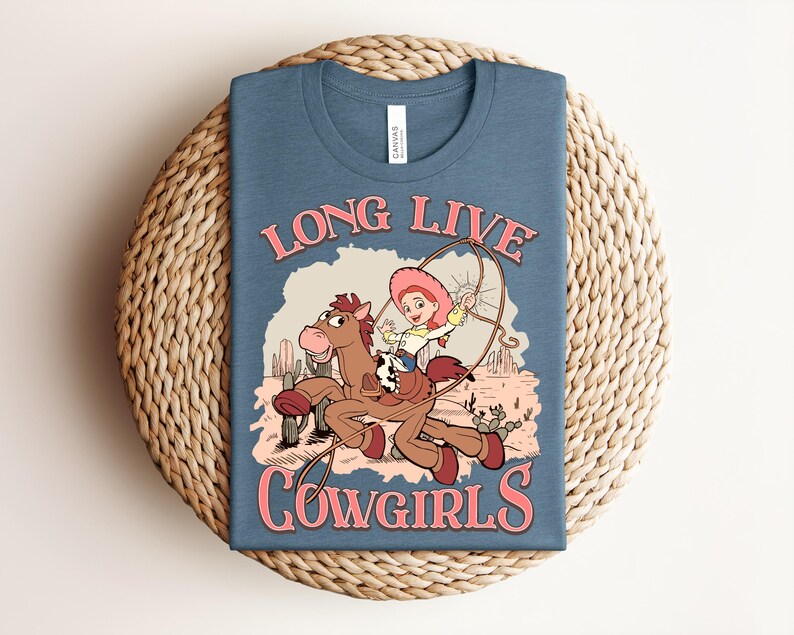 May include: A blue t-shirt featuring a graphic of a cartoon cowgirl riding a brown horse while holding a lasso. The text "LONG LIVE COWGIRLS" is printed in pink above and below the illustration, which includes cacti and a desert landscape.