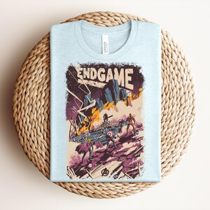Pu&ograve; includere: Una t-shirt azzurro chiaro con una stampa grafica che riporta il testo ENDGAME in alto. L'illustrazione raffigura una scena di battaglia con figure stilizzate, fiamme arancioni, terreno roccioso viola e fulmini, con il logo Avengers in basso.