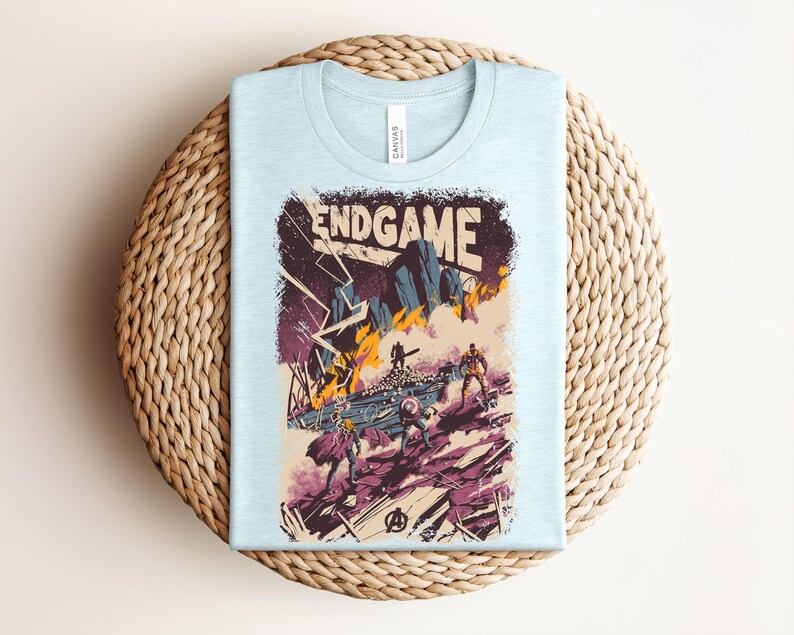 Pu&ograve; includere: Una t-shirt azzurro chiaro con una stampa grafica che riporta il testo ENDGAME in alto. L'illustrazione raffigura una scena di battaglia con figure stilizzate, fiamme arancioni, terreno roccioso viola e fulmini, con il logo Avengers in basso.