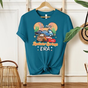 K&ouml;nnte beinhalten: T&uuml;rkisfarbenes T-Shirt mit einer Grafik von Zeichentrickautos aus dem Film Cars und dem Text "In my Radiator Springs ERA". Das Design umfasst Kakteen und eine sonnen&auml;hnliche Form. Das T-Shirt ist unten geknotet.