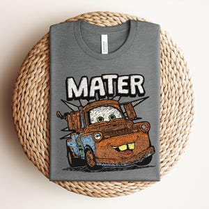 May include: A heather gray t-shirt featuring a graphic print of a rusty brown tow truck character with green eyes and a wide smile. The word MATER is printed in large white letters above the truck. The design includes a black and white checkered burst.