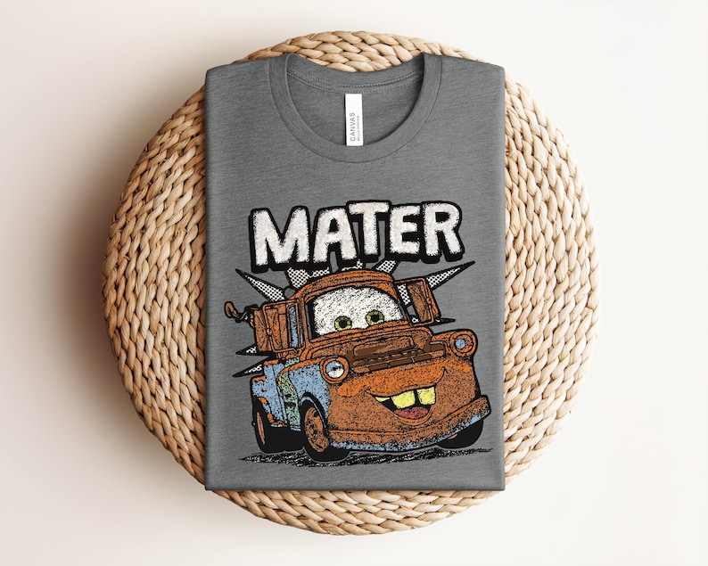 May include: A heather gray t-shirt featuring a graphic print of a rusty brown tow truck character with green eyes and a wide smile. The word MATER is printed in large white letters above the truck. The design includes a black and white checkered burst.