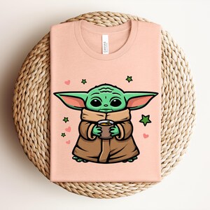 May include: A light peach-colored t-shirt featuring a cartoon illustration of a green alien character holding a cup. The character is wearing a brown cloak and has large ears. Pink hearts and green stars surround the character.